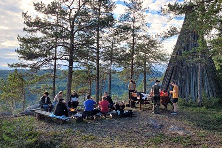 En grupp unga människor sitter runt en brasa på en höjd i en skog intill ett vindskydd i trä.