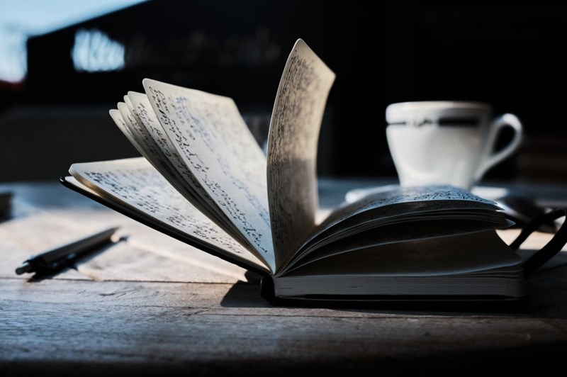 Bok som vänder blad. Mörk bakgrund och en kaffekopp finns bredvid boken. Boken ligger på ett bord och bredvid boken finns en penna.