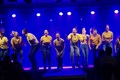 En ensemble klädd i jeans och gula tröjor står på en scen och sjunger. 