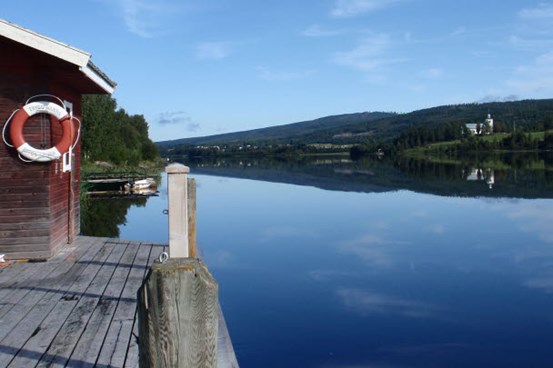 En brygga med en sjöbod bredvid den spegelblanka Ångermanälven och en blå himmel.
