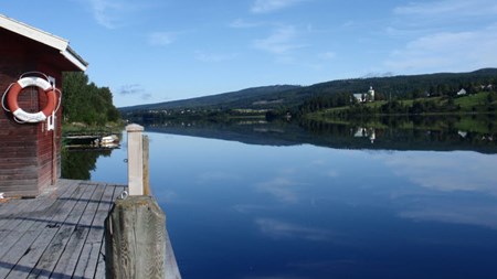 En brygga med en sjöbod bredvid den spegelblanka Ångermanälven och en blå himmel.