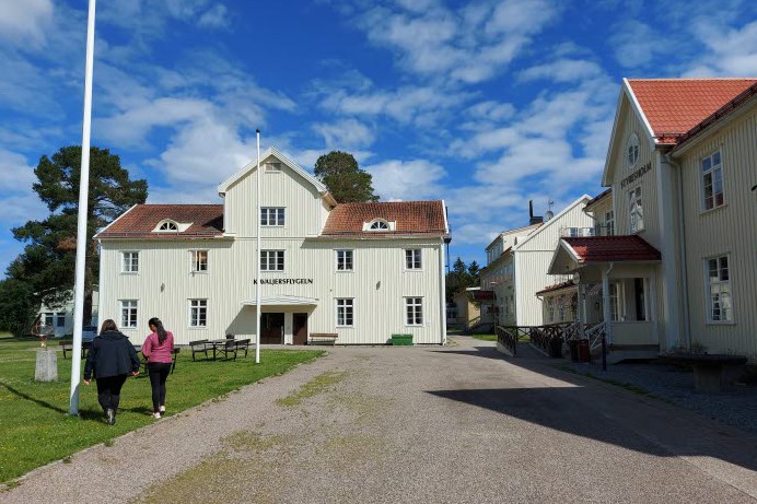 Hola folkhögskolas ena skolbyggnad, en blå himmel och två personer som går över en grusplan framför byggnaden.