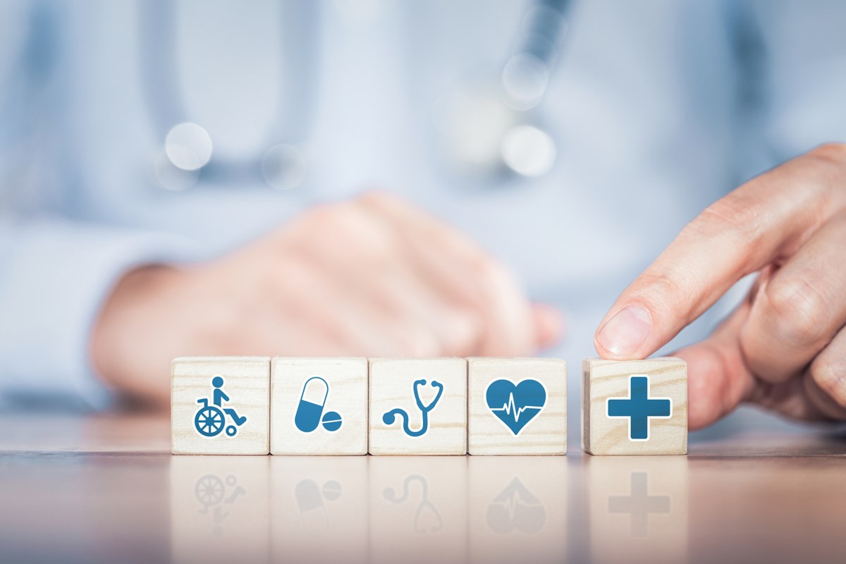 alt='En hand lägger klossar på ett bord. Klossarna har symboler för hälsa och sjukvård: En rullstol, mediciner, ett stetoskop, ett hjärta och ett  plus.'