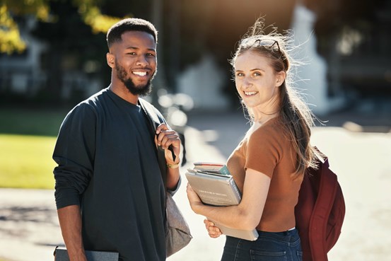 En manlig och en kvinnlig student står och håller i skolböcker utomhus och tittar glatt in i kameran.