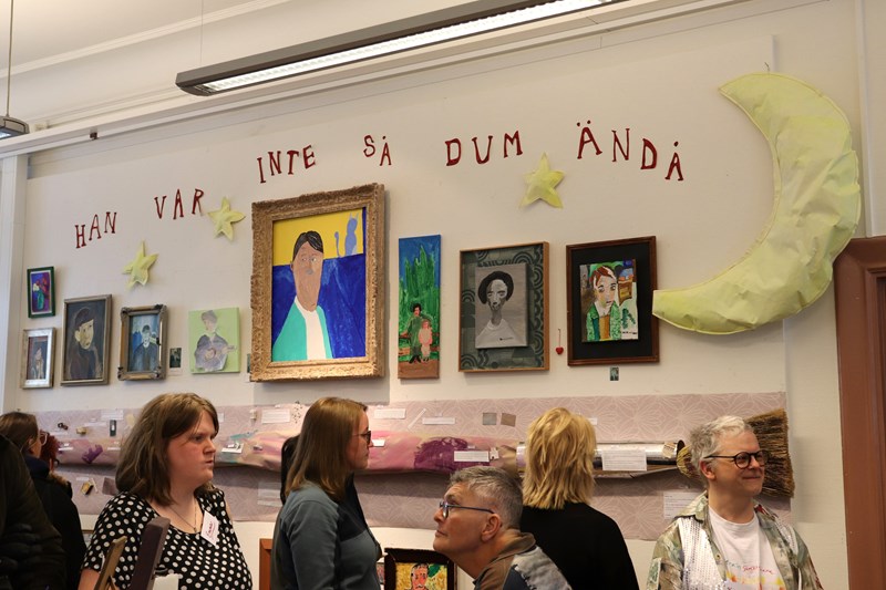 En vägg full av målningar och människor som minglar under en vernissage.