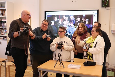 En pedagog står tillsammans med en grupp glada deltagare och visar något på en systemkamera.