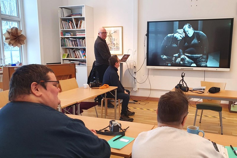 En pedagog står längst fram i ett klassrum och visar foton på en stor bildskärm framför tre deltagare som sitter vid skolbänkar.