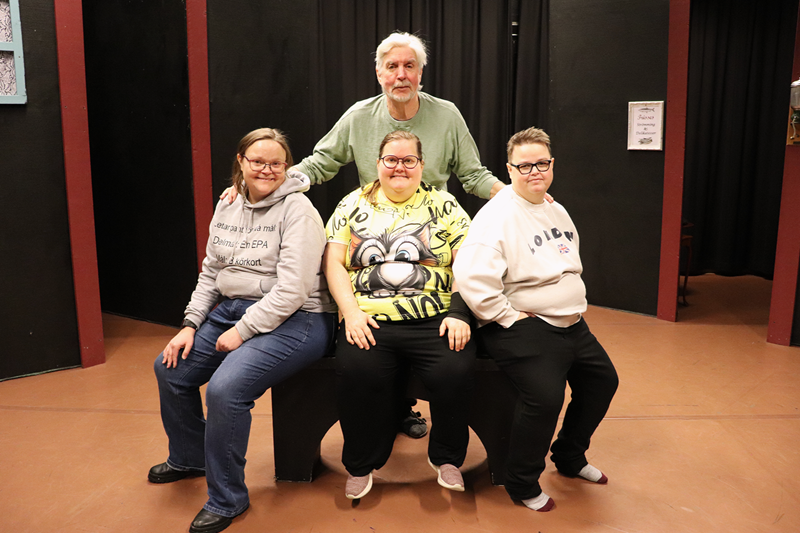 En pedagog står bakom tre deltagare som sitter på en bänk i en teatersal..