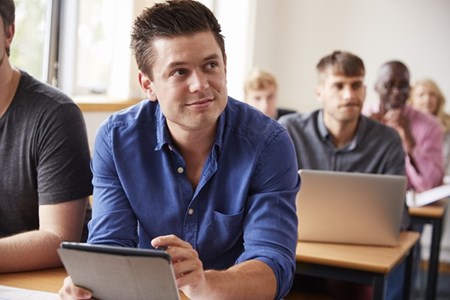 En man sitter med en surfplatta i ett klassrum tillsammans med några kurskamrater.