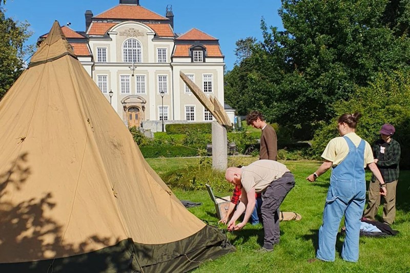 En grupp deltagare sätter tillsammans upp en tältkåta i parken med skolans huvudbyggnad i bakgrunden.