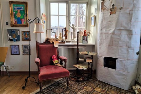 En gammal fåtölj och en lampa står bredvid en kakelugn gjord i papper och i bakgrunden hänger färgstarka tavlor