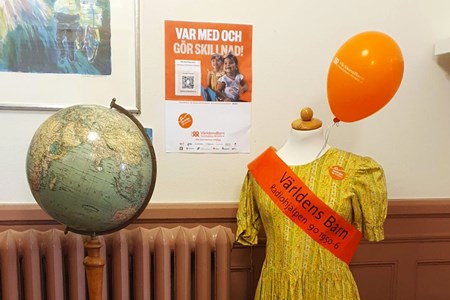 En provdocka i gul klänning och orange banderoll står bredvid en jordglob, en orange ballong och en affisch för Världens Barn.