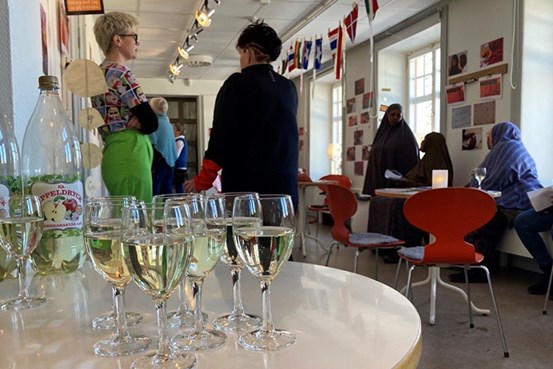 I förgrunden står vinglas fyllda med äppelcider med en grupp personer står och sitter i bakgrunden