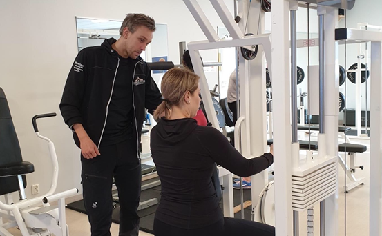 En kvinna demonstrerar för en man hur en av gymmets maskiner fungerar.