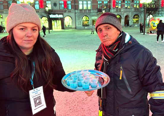 En fluortant står med en bricka med fluoridlösning och en besökare står bredvid och sköljer, utomhus på torget i Sundsvall.