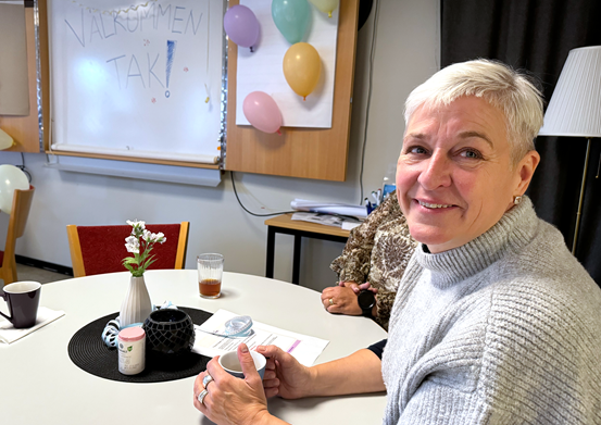 Kvinna sitter vid ett fikabord och tittar in i kameran.