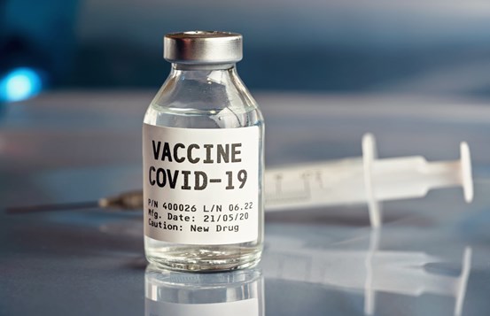 Närbild på flaska med text "vaccin covid-19"  och i bakgrunden en spruta