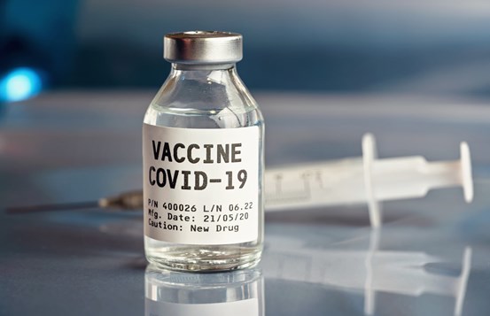 Närbild på flaska med text "vaccin covid-19" och i bakgrunden en spruta