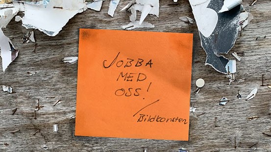 En orange papperslapp med texten "Jobba med oss! /Bildkonsten". Papperslappen ser ut att sitta på en välanvänd anslagstavla med delar av gamla anslag och häftor. 