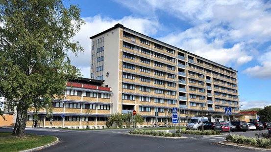 Sjukhuset i Sollefteå, en byggnad på flera våningar. I förgrunden asfalterad gata, i bakgrunden en blå himmel men även moln. Till vänster ett grönt träd.