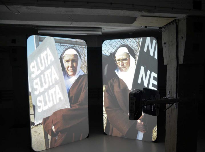 På två stående skärmar visas stillbilder från ett videoverk. Här syns två nunnor med plakat där texten ”SLUTA SLUTA SLUTA” står i versaler, samt delar av ett annat plakat med delvis avklippt text som dock kan tolkas som ”NEJ NEJ”. 