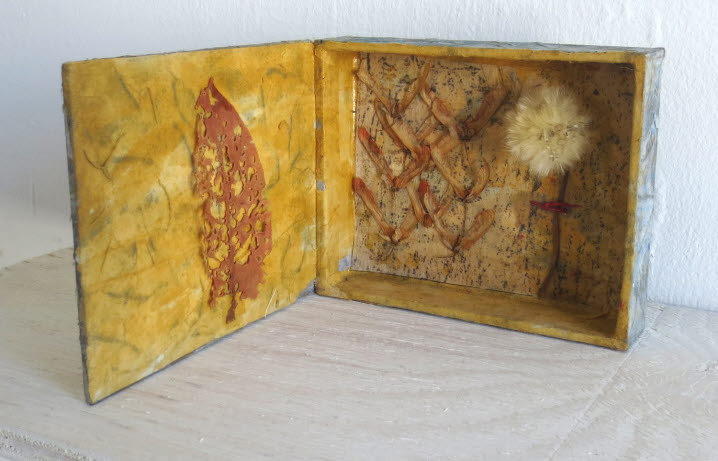 Objekt av Christer Carlstedt. Skåp med locket öppet, insidan gul. Ett rött löv på insidan av locket och en fröfjunig maskros i skåpet.