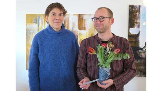 Fotografi av 2019 års Härkestipendiat Kristian Berglund tillsammans Anna Erlandsson från stipendegruppen. Stipendiaten håller blommor i handen och bakom dem skymtar två målningar.
