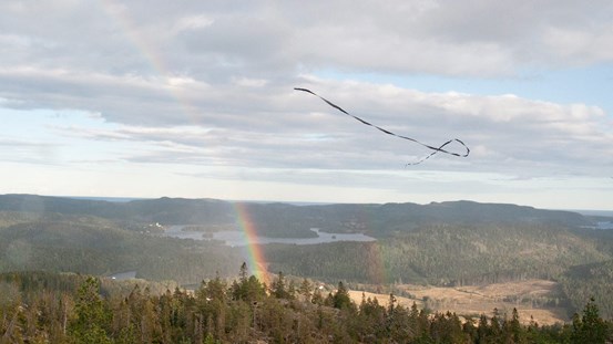 En svävande drake över ett vidsträckt landskap med både blå och molnig himmel. En regnbåge syns till vänster. 