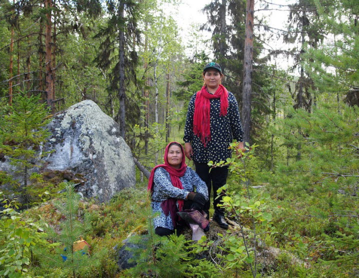 Två personer står, respektive sitter, på en sten i skogen, med röda sjalar och bärplockare. 