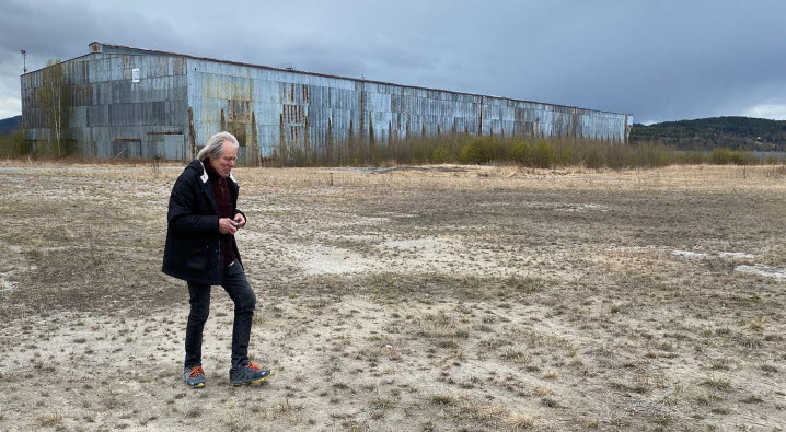 Över ett övergivet industriområde traskar Janne Björkman med kamera i hand och blicken vänd mot marken. I bakgrunden syns en stor plåtbyggnad och en liten del av älven skymtar.