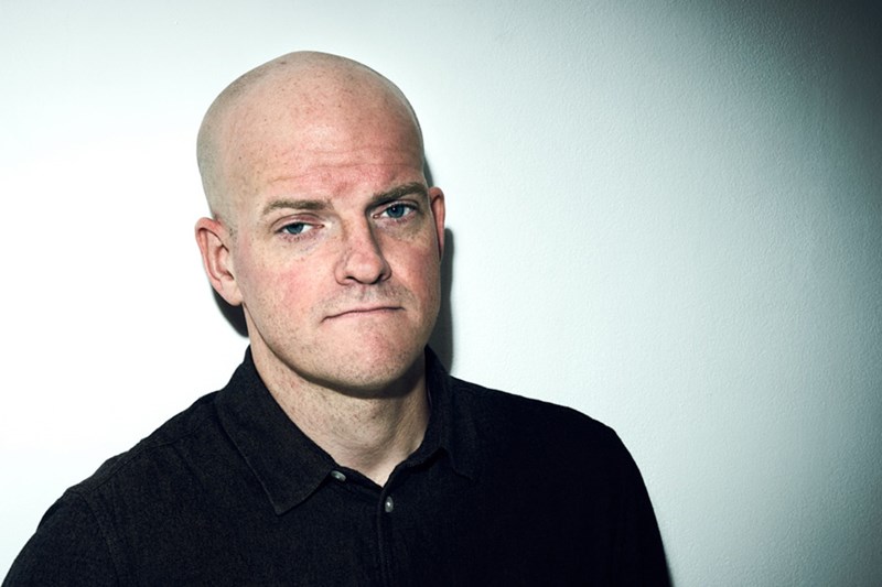 Foto. Porträtt av Henrik Bromander, med blicken mot betraktaren, i mörk skjorta och vit bakgrund.